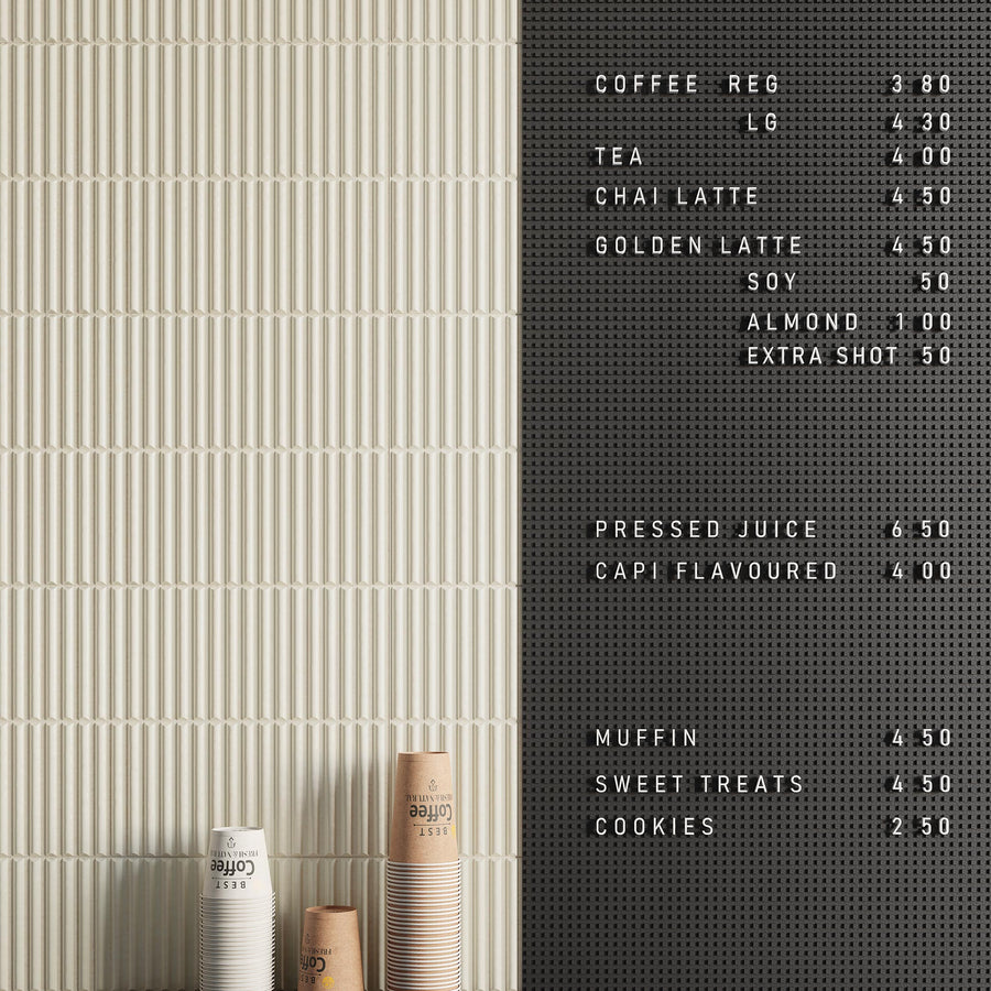 A café menu board lists drinks and prices on a black pegboard wall. Three stacks of disposable coffee cups sit on a patterned beige counter below the menu. The wall is half beige tiles, half black pegboard.