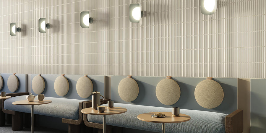 Modern minimalist cafe interior with light-colored cushioned bench seating, round pillows, small tables, mugs, plates, and wall-mounted lights on a textured cream wall. The atmosphere appears calm and inviting.
