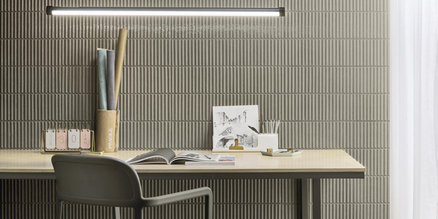 A modern desk with an open book, pens, a notebook, and art prints sits against a ribbed gray wall under a slim, horizontal light fixture. A black chair is in front of the desk, and light filters through a sheer curtain.