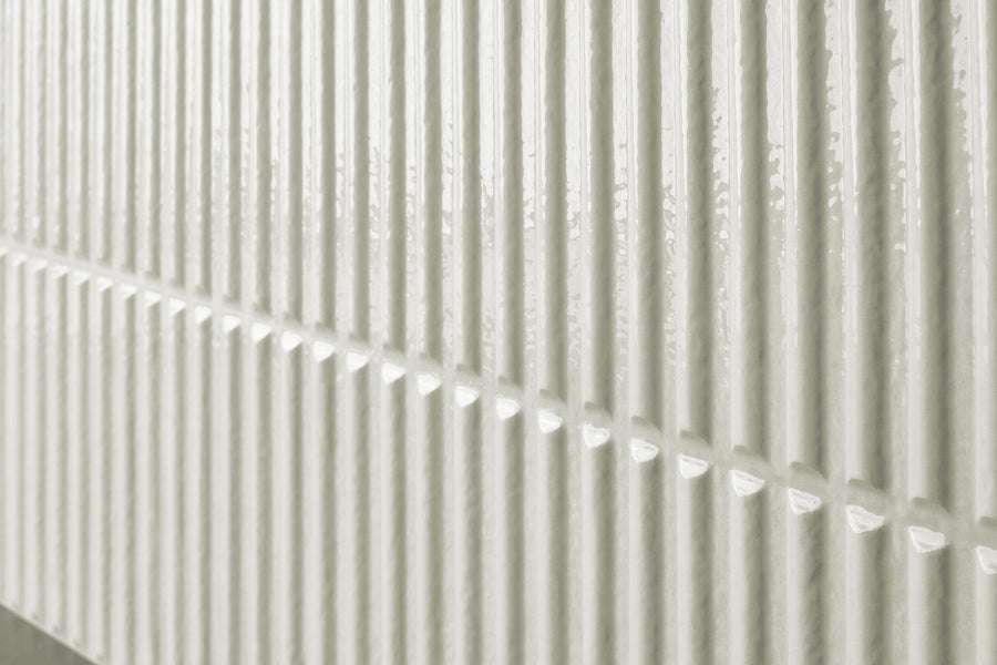 Close-up of a wall with vertical rectangular white ceramic tiles, featuring a glossy finish and a raised, textured surface that creates a repeating linear pattern.