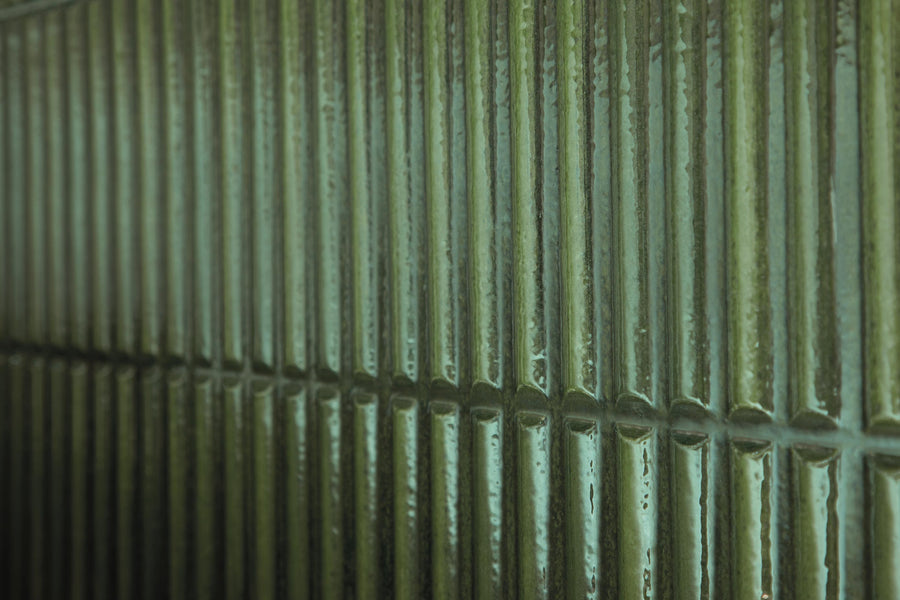 Close-up of a wall covered with glossy, rectangular, dark green tiles arranged in a vertical pattern, with reflections of light creating a textured and shiny appearance.