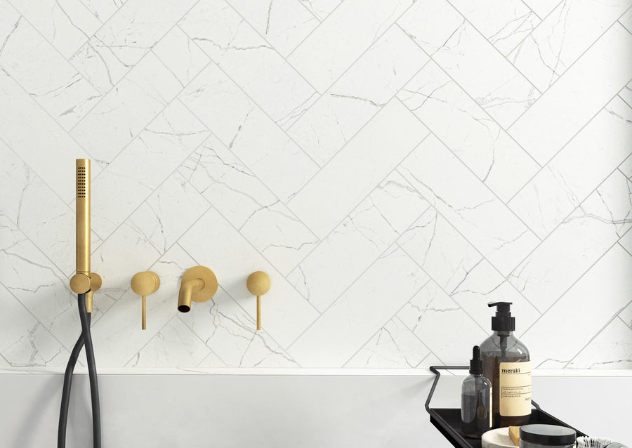 Modern bathroom with white herringbone-patterned marble tiles, matte gold shower fixtures, and a black shelf holding soap, bottles, and a jar. The style is minimal and elegant.