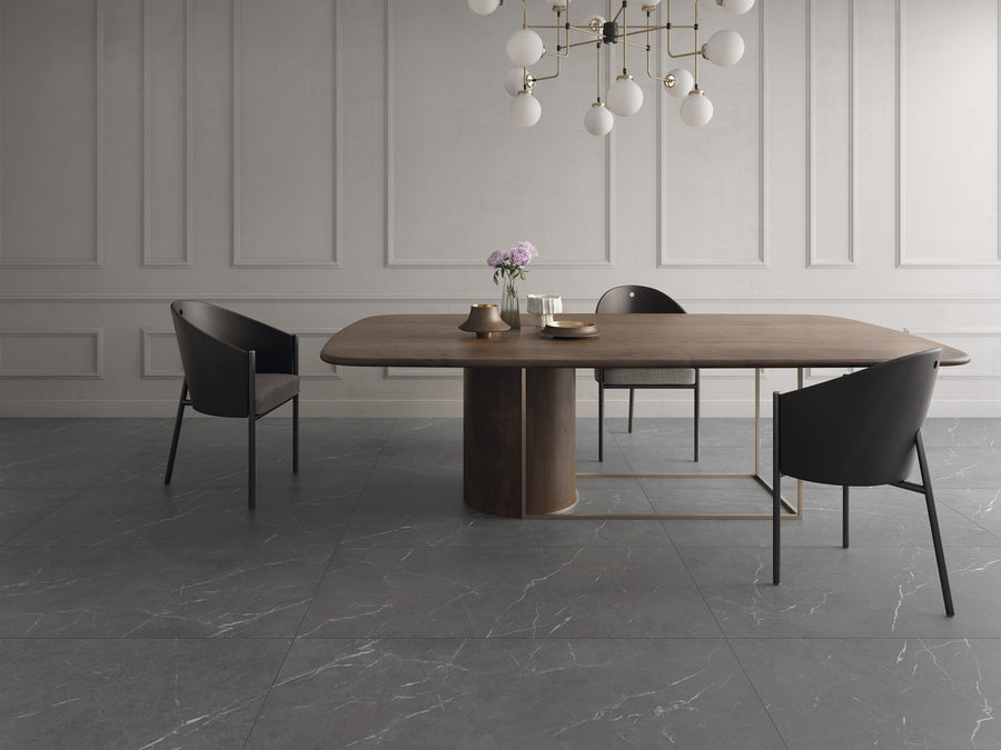 Modern dining room with a rectangular wooden table, two black chairs, a gold chandelier with globe lights, and a vase of flowers on the table. The room has gray marble flooring and white paneled walls.