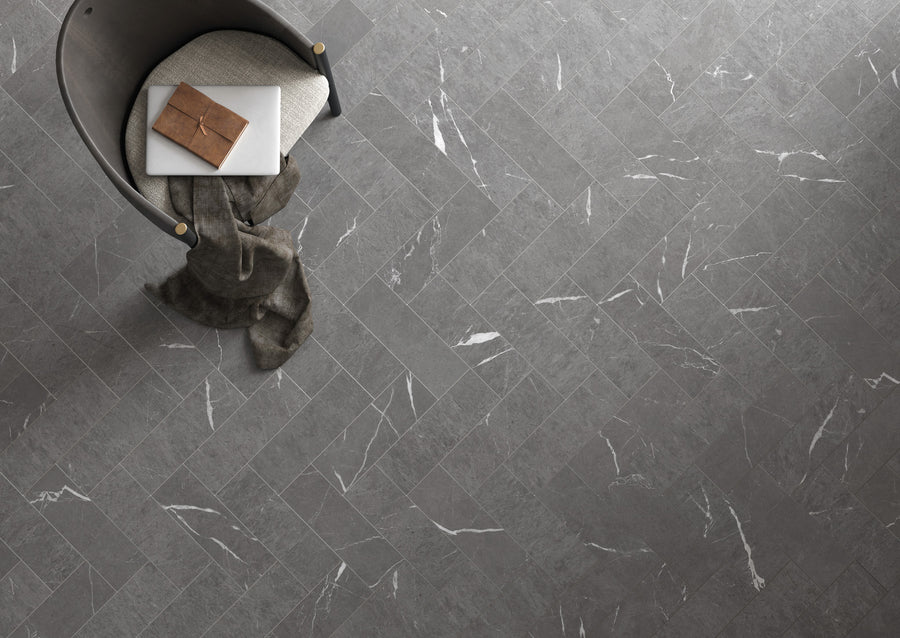 A top-down view of a gray herringbone-patterned tile floor with white veining. In the corner, theres a modern chair with a cushion, a brown notebook, a white tablet, and a draped gray throw blanket.