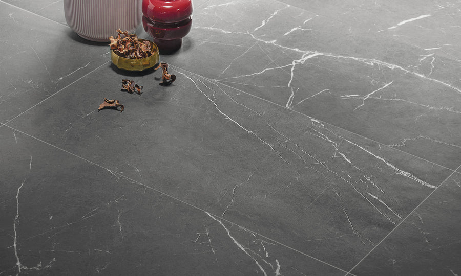 Close-up of large, dark grey marble tiles with white veining. On the floor, there is a ribbed white vase, a red candle holder, and a small green bowl filled with dried brown leaves.