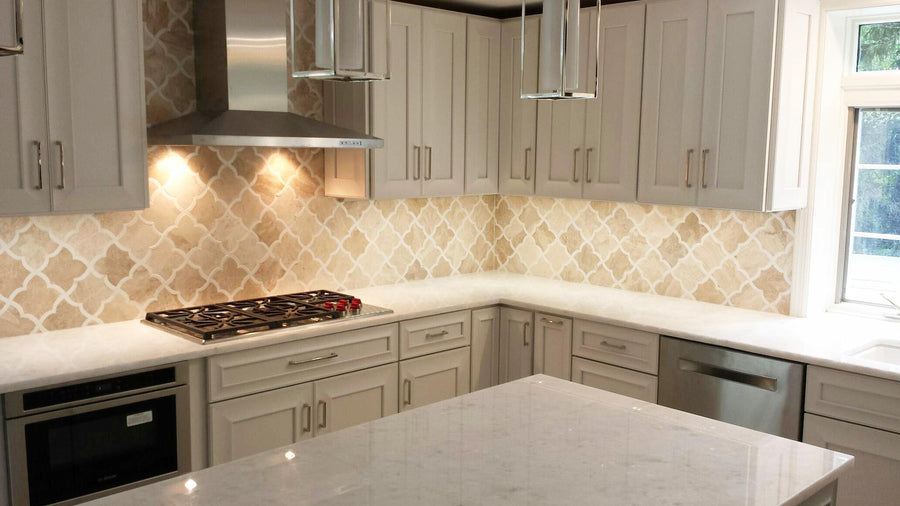 Modern kitchen with white cabinets, a patterned beige tile backsplash, stainless steel stove, oven, dishwasher, and hood, white marble countertops, and pendant lights above a central island.