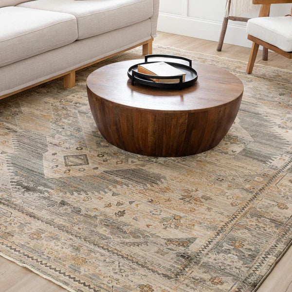 A round, wooden coffee table with a black tray on top sits on a large, patterned beige and gray rug in a living room, next to a light-colored sofa and a wooden chair.