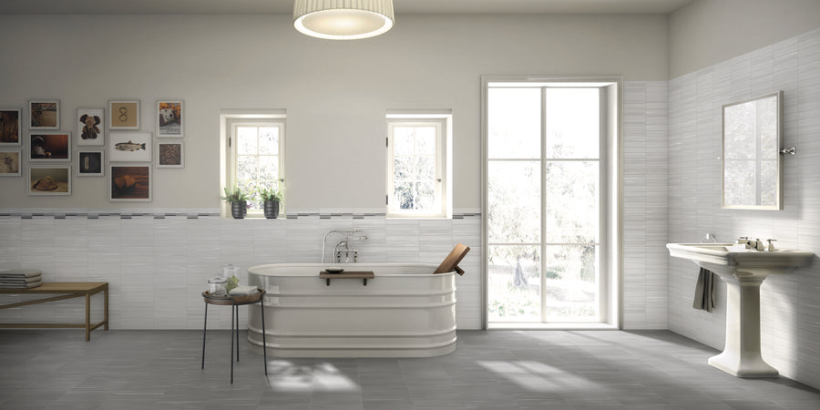 A bright, modern bathroom with a freestanding white bathtub, pedestal sink, and large window. Art and plants decorate the walls, and gray tile flooring complements the light-colored walls and fixtures.