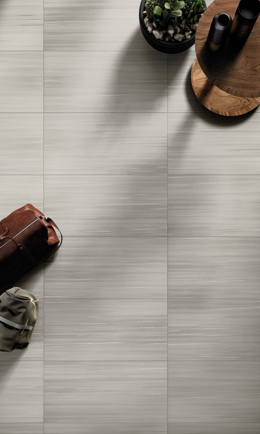 A top-down view of a light grey, textured tile floor with a brown leather bag, a folded khaki garment, a round wooden table, and a black potted plant on the surface.