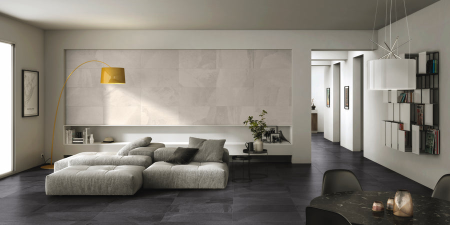 Modern living room with a light gray sectional sofa, black coffee table, and a yellow floor lamp. The space features dark tile flooring, minimal decor, and built-in shelving with plants and art pieces.