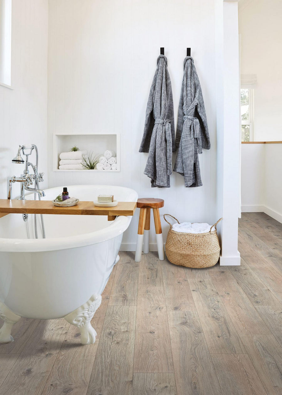 A modern bathroom with a white clawfoot bathtub, wood plank floor, two gray robes hanging on hooks, a wicker basket with towels, and a wooden stool beside the tub. Towels and toiletries are neatly arranged on shelves.