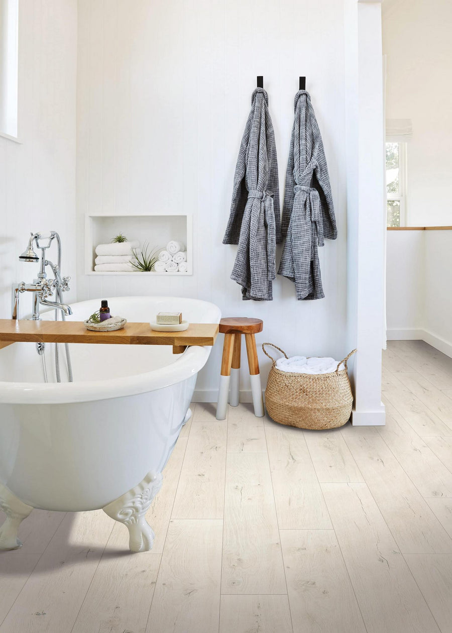 Bright, modern bathroom with a white clawfoot tub, wooden bath tray, two gray robes hanging on the wall, a small wooden stool, rolled towels, and a woven laundry basket on light wood flooring.
