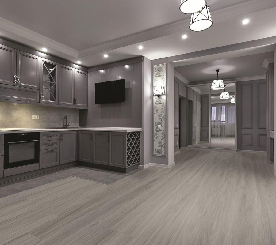 Modern kitchen and dining area with gray cabinets, built-in oven, and wall-mounted TV. Light wood flooring, recessed and pendant lights, and an open layout leading to additional rooms and large windows.