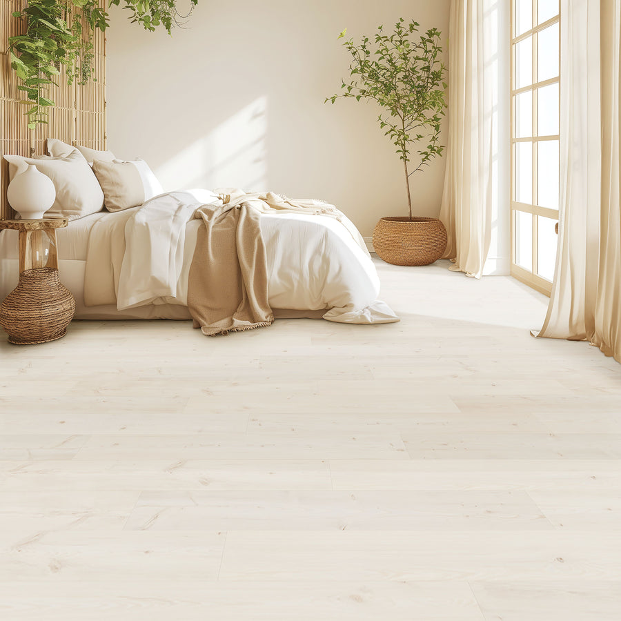 A bright, minimalist bedroom features Glacial Isle 8 11/32" Waterproof Laminate in light wood tones, a cozy bed with white and beige bedding, wicker accents, potted plants, and large windows letting in natural sunlight.