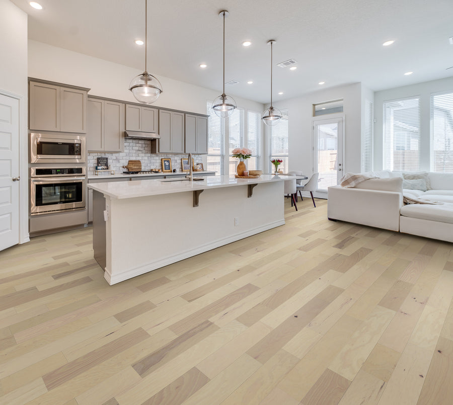 Modern kitchen with Deepwood Trail 5″ Hickory Oak Hardwood floors, beige cabinets, white island with pendant lights above, and an open living area featuring a white sectional sofa and large windows letting in natural light.
