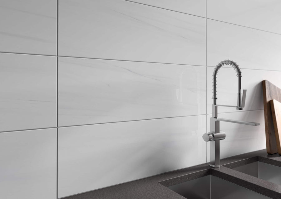 A modern kitchen sink with a sleek, stainless steel faucet is set against a wall of large, glossy white tiles. A wooden cutting board rests on the dark countertop next to the sink.