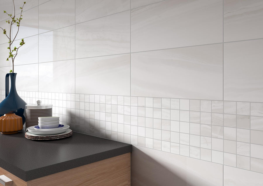 Modern kitchen counter with stacked plates, bowls, a blue vase with green branches, and a small orange container. The wall features large white tiles and a decorative strip of small square tiles.