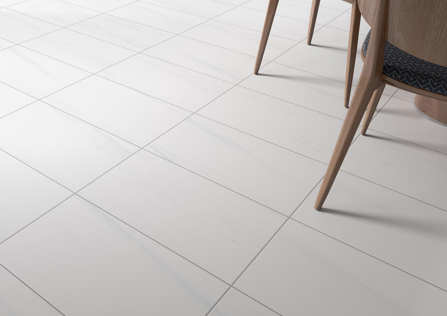 Close-up view of a floor with large white ceramic tiles featuring subtle gray veining. The legs of a wooden table and chairs with upholstered seats are visible in the upper right corner.