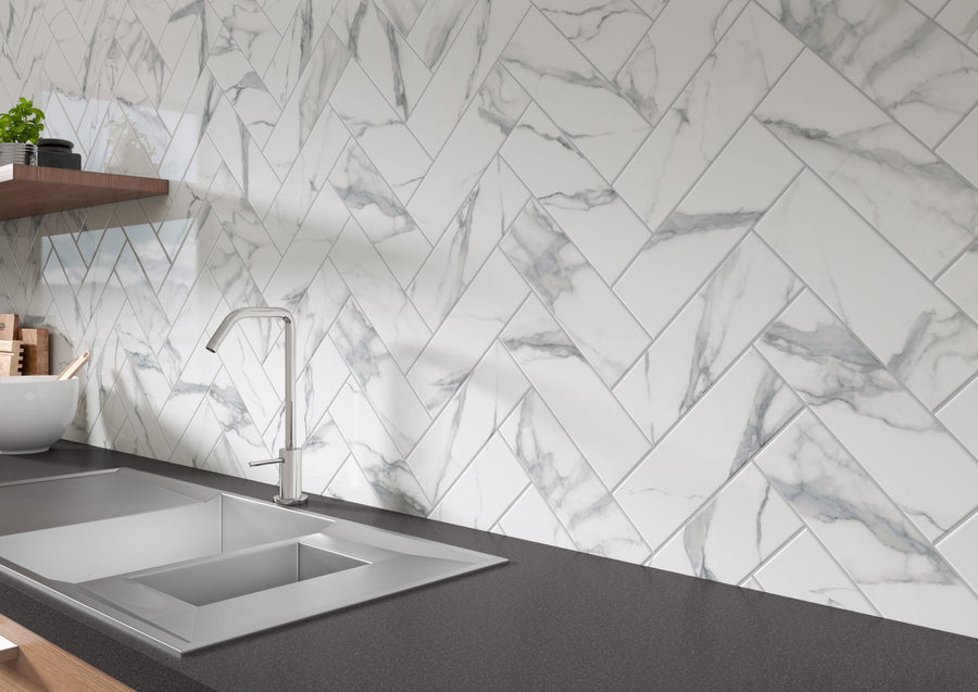 A modern kitchen countertop with a stainless steel sink and faucet, dark surface, and a backsplash of white marble tiles arranged in a herringbone pattern. Wooden shelves with bowls and plants are visible in the background.