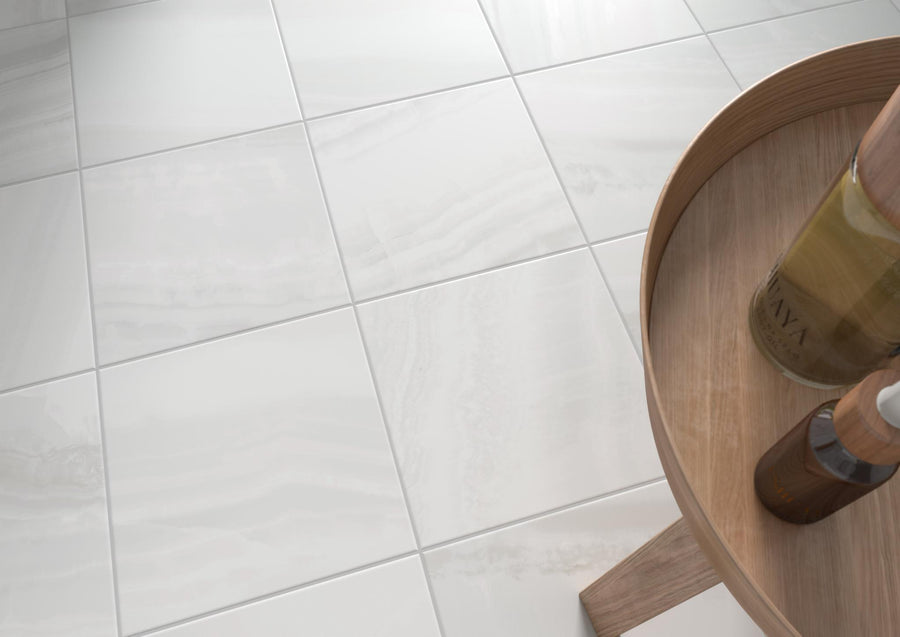 Close-up of glossy white ceramic floor tiles with subtle gray veining. Part of a round wooden side table with bottles is visible in the lower right corner.