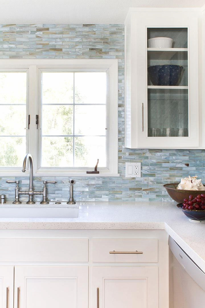 Bright kitchen with white cabinets and countertop, a stainless steel sink under a window, blue mosaic tile backsplash, a glass-front cabinet, and bowls of fruit and shells on the counter.