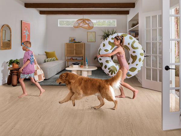 Two children in swimsuits and goggles, one holding a striped bag and the other carrying a large pool float, walk through a bright living room with a golden retriever. The room has modern decor and natural light.