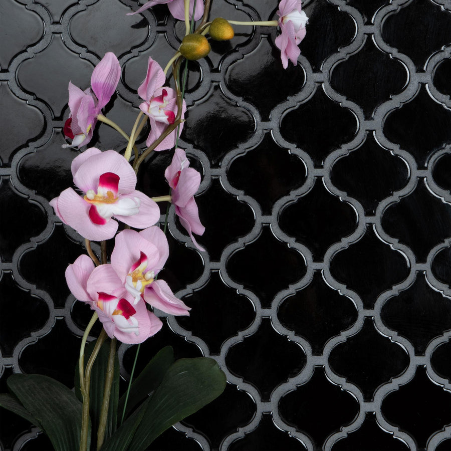A stem of pink orchids with green leaves is positioned against a glossy black tile wall featuring an ornate, repeating geometric pattern.