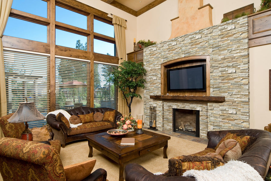 A spacious living room with large windows, a stone fireplace, a mounted TV, brown leather sofas with cushions, a wooden coffee table, and decorative plants and lamps. Natural light fills the cozy space.