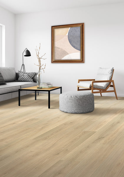 A modern living room with light wood flooring, a gray sofa, black coffee table, light gray ottoman, cushioned wooden armchair, abstract wall art, and a floor lamp. The decor is minimalist with neutral colors.