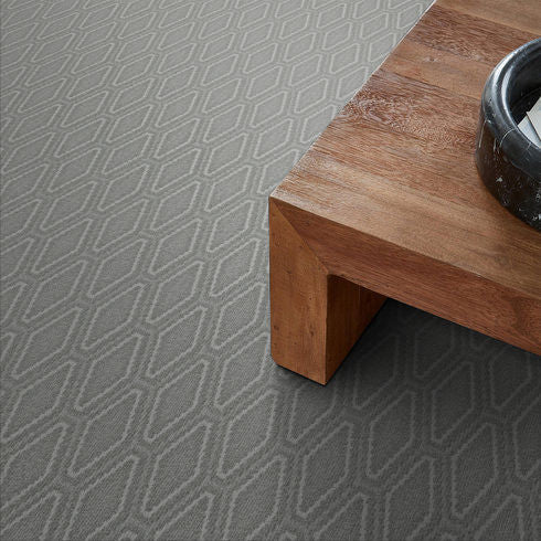 A close-up view of a gray carpet with a repeating diamond pattern next to the corner of a wooden table with a black round decorative object on top.