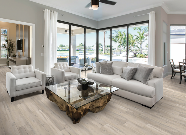 Modern living room with light-colored sofas and armchairs, a glass coffee table with a wooden base, large windows showcasing a patio, palm trees, and a pool outside, and neutral-toned decor throughout.