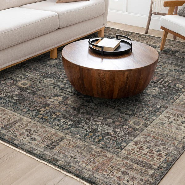 A round wooden coffee table sits on an ornate, patterned area rug in a cozy living room with a beige sofa and a wooden armchair visible in the background.