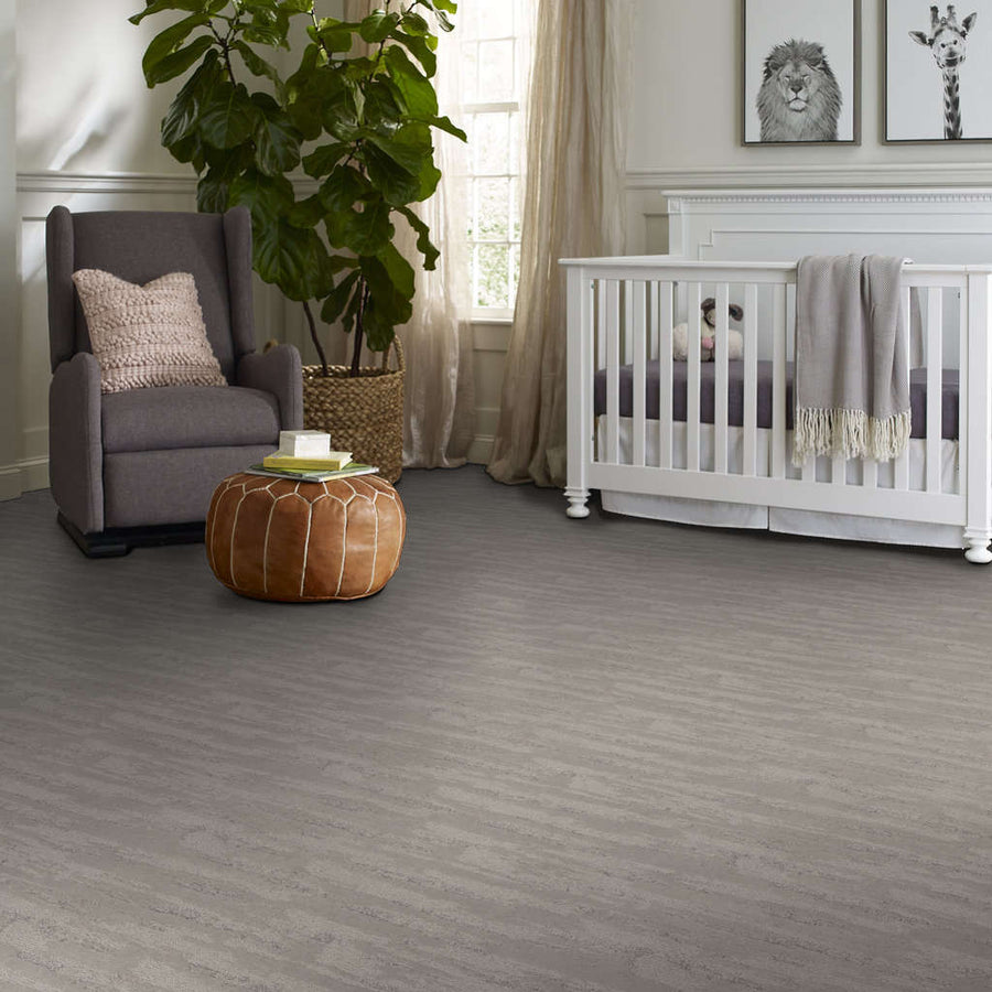 A cozy nursery with a gray armchair, leather pouf, large plant, white crib with a gray blanket, and animal artwork on the wall, all on a gray wood-patterned floor. Light streams in from a window.