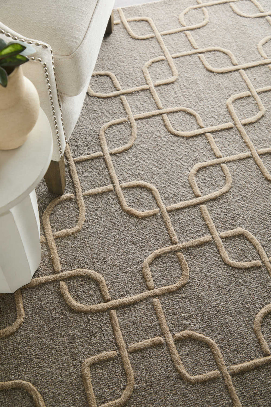 A beige armchair next to a white round table, placed on a textured carpet with an interlocking geometric pattern in shades of brown and tan.