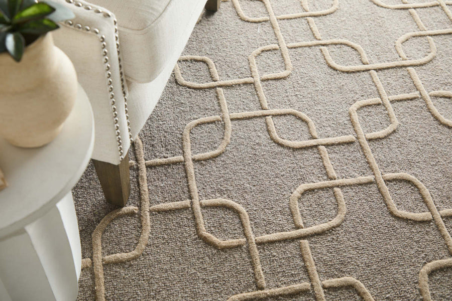 A close-up of a beige and taupe textured rug with a raised geometric pattern, next to a cream-colored upholstered chair and a white round table with a potted plant.