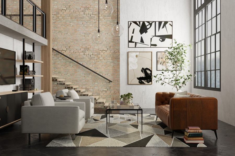 A modern living room with a brick accent wall, large windows, gray armchairs, a brown leather sofa, geometric rug, glass coffee table, and abstract art on the walls. A staircase is visible in the background.
