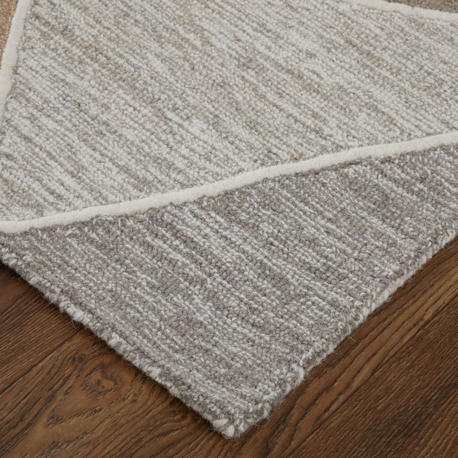 A close-up of the corner of a textured, light gray and cream rug with diagonal lines, placed on a medium brown wooden floor.