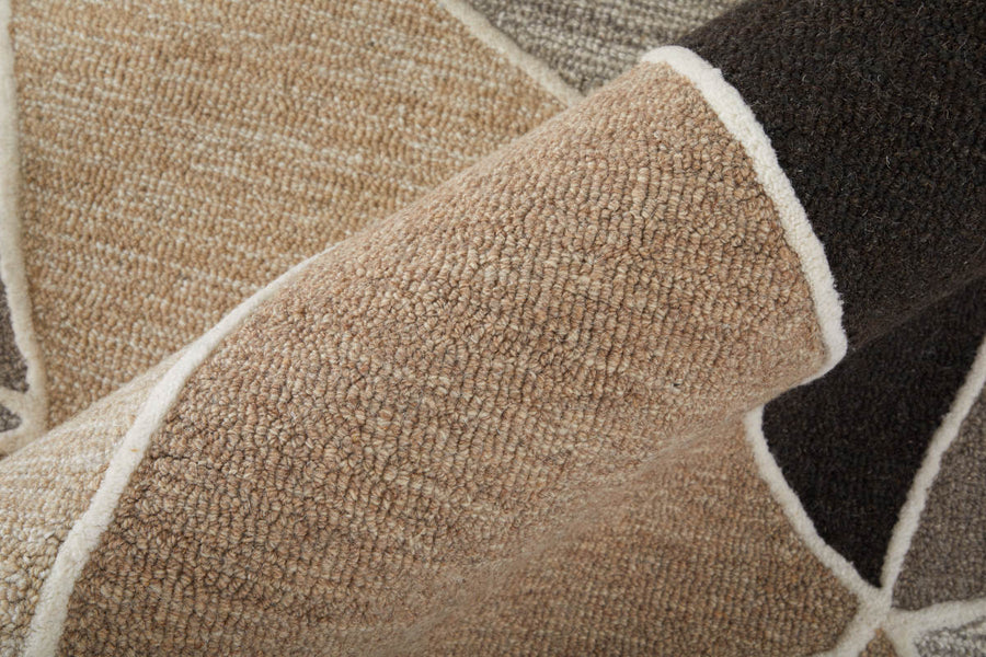 Close-up view of a textured, woven rug with geometric patterns in beige, brown, and gray tones, bordered by cream-colored lines. The rug is folded, highlighting its thickness and texture.
