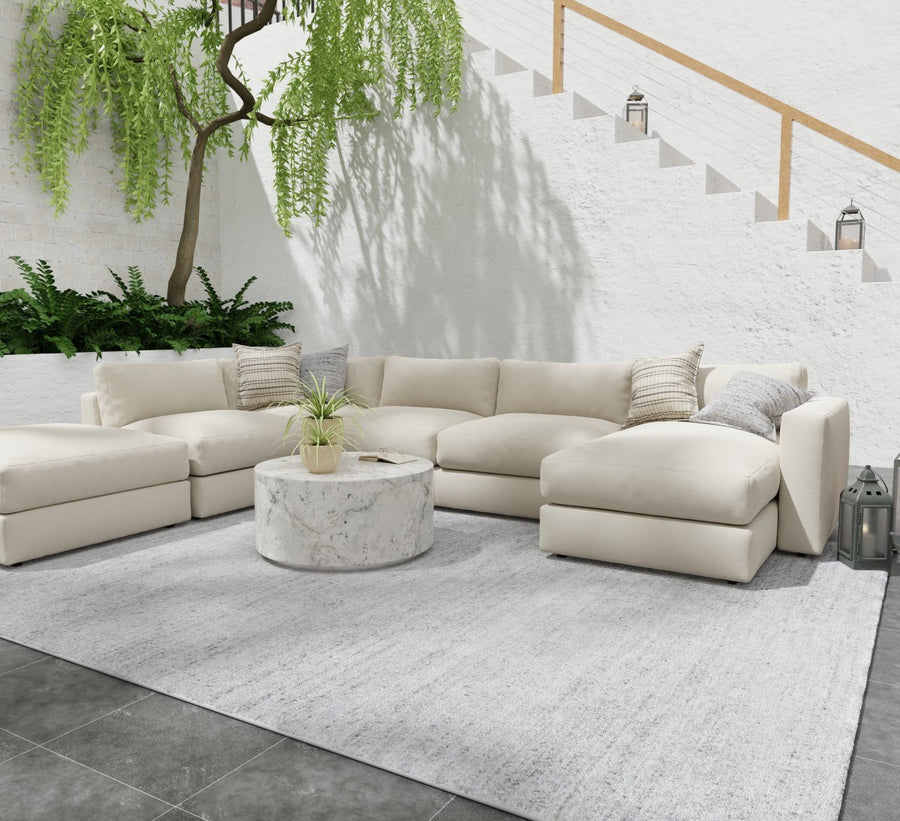 A modern outdoor patio with a cream sectional sofa, marble round coffee table, light gray rug, and potted plants. White stairs and a leafy green tree are in the background against a textured white wall.