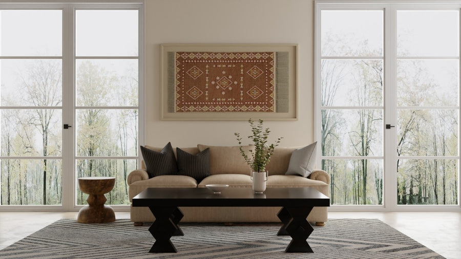 A modern living room features a beige sofa with dark pillows, a black coffee table, a small wooden side table, a patterned rug, a vase with greenery, and large windows overlooking trees. A decorative tapestry hangs on the wall.