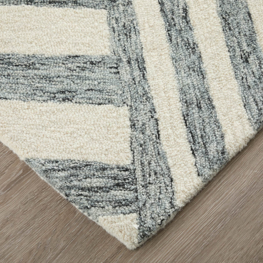 A close-up view of a rug corner with a geometric pattern in cream and gray tones, placed on a light wood floor. The rug features textured stripes in alternating colors.