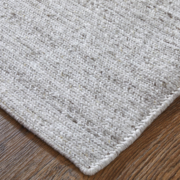 Close-up of a light gray, woven textured area rug with subtle specks of darker fibers, placed on a wooden floor. The rug has a rounded, finished edge visible in the foreground.