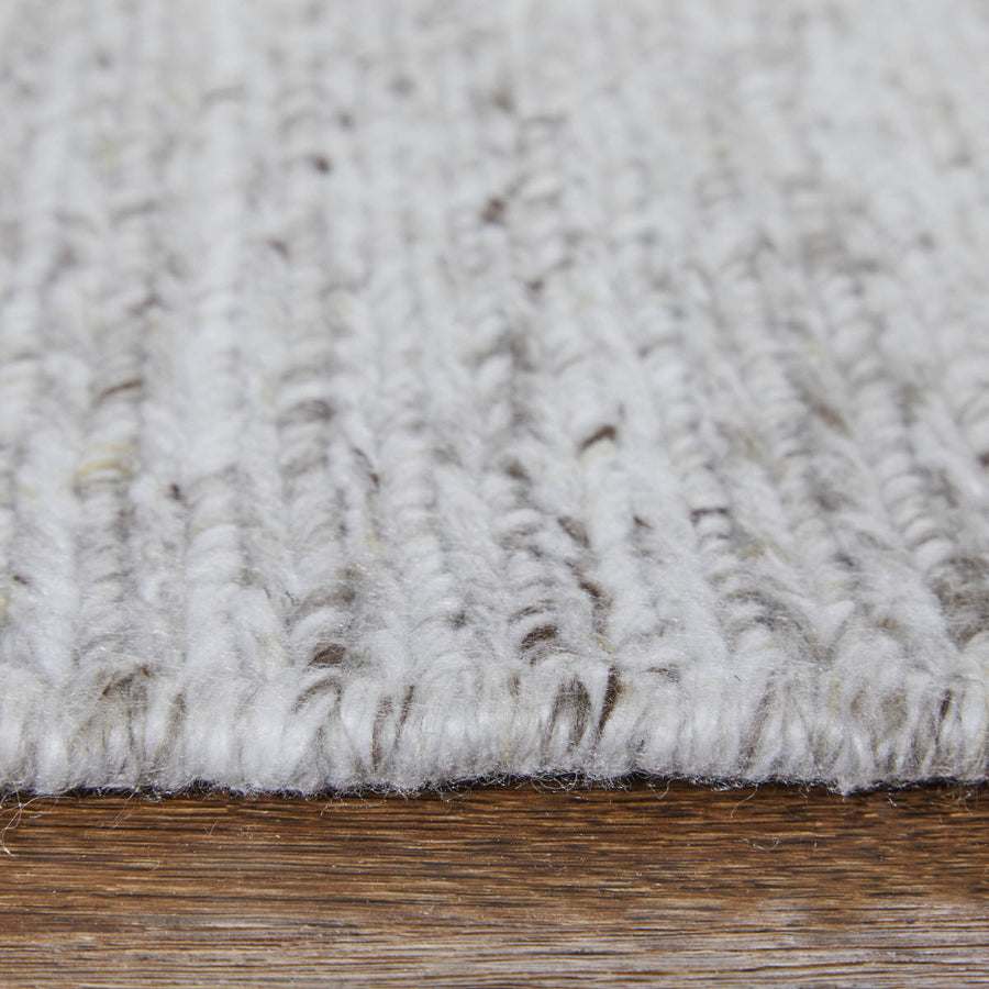 Close-up of a textured, light grey woven rug with thick yarns, placed on a wooden floor. The image highlights the rug’s fibers and surface pattern.