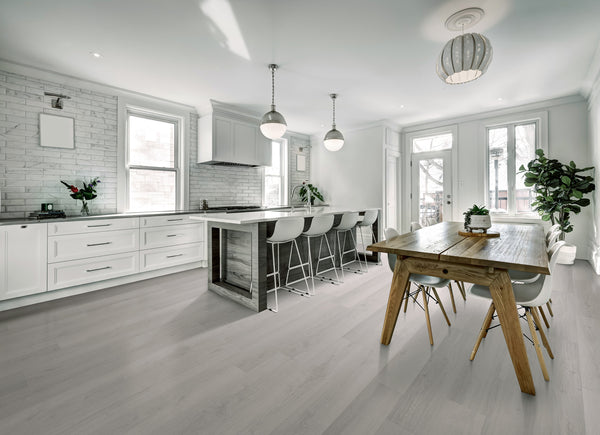 Modern kitchen and dining area with white cabinets, a large island with barstools, wooden dining table with chairs, pendant lighting, and large windows letting in natural light; plants add a touch of greenery.