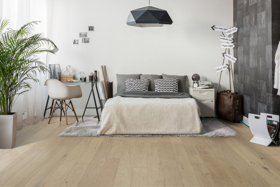 A modern bedroom with a cozy bed, gray and white decor, a desk with a chair, wall photos, a potted plant, and a signpost decoration beside luggage near a textured accent wall. Natural light fills the room.