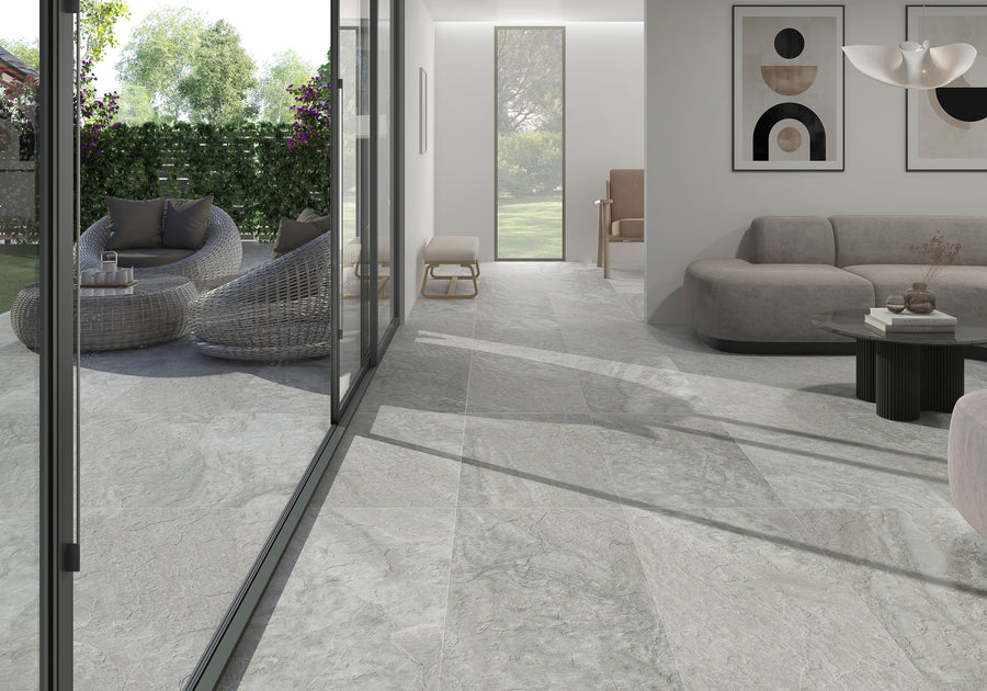 Modern living room with large gray marble floor tiles, light gray sofa, abstract wall art, and glass sliding doors leading to a patio with wicker chairs and a garden view. Bright natural light fills the space.