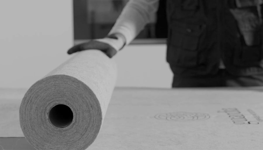 A person in a dark vest and glove is unrolling a large roll of Guardia Surface Protection onto a flat surface, likely preparing it for cutting or installation. The black-and-white image highlights the product’s waterproof feature.