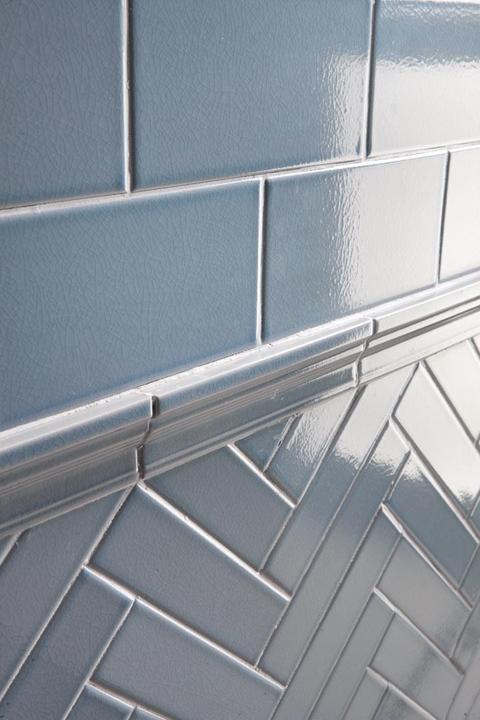 Close-up of a wall with glossy light blue tiles; the upper section has rectangular tiles laid horizontally, separated by a trim, while the lower section features tiles arranged in a herringbone pattern.