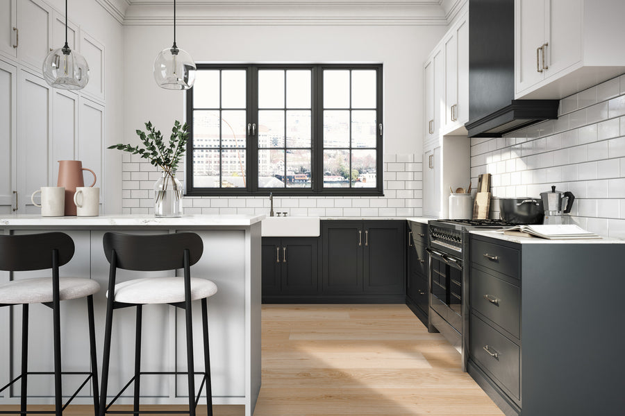 Modern kitchen with black and white cabinets, a large window above the sink, two black barstools at a white island, pendant lights, light wood flooring, and a vase with green branches on the countertop.