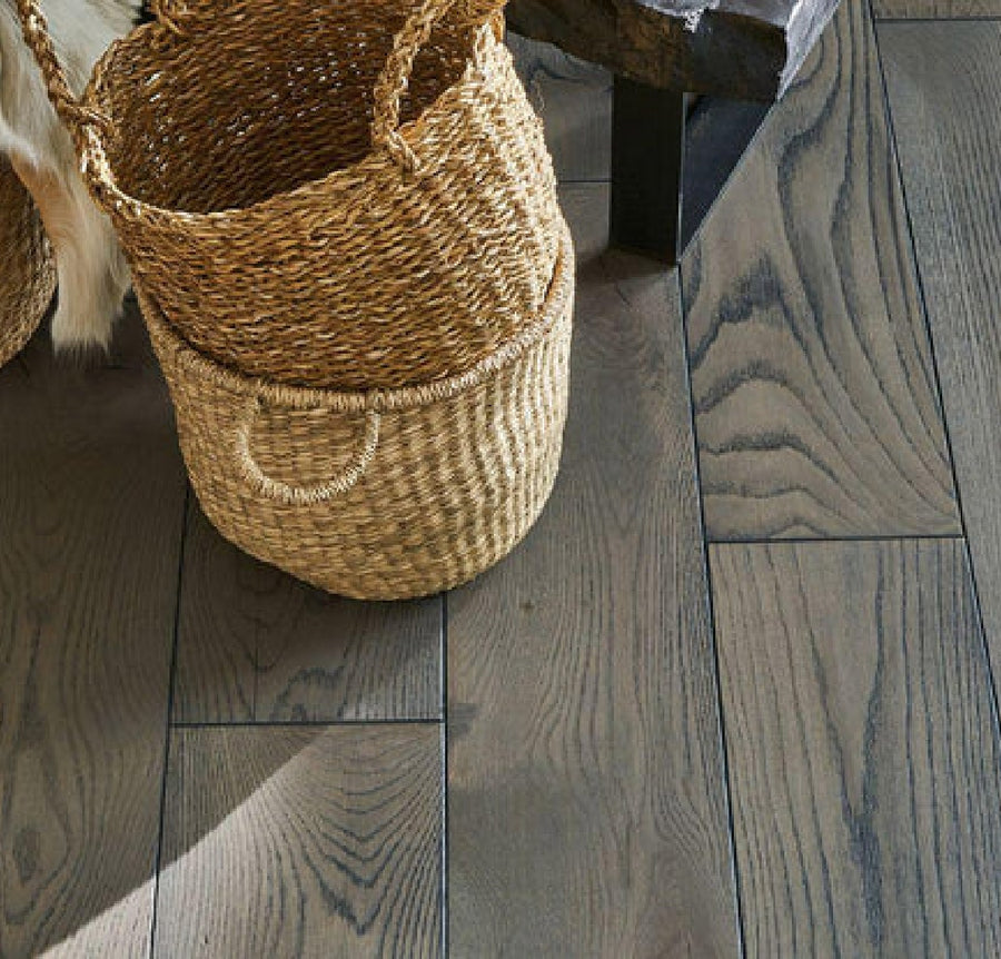 Two woven baskets with handles sit on a floor made of Commercial Hardwood next to dark wooden furniture. A portion of light-colored animal hide is visible at the edge of the image.